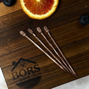 May include: Four copper cocktail stirrers with a twisted design and a heart-shaped end. The stirrers are arranged on a wooden surface with a logo that reads "BORS DESIGN CO."