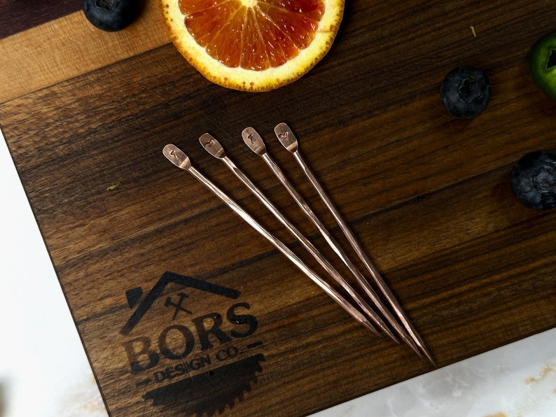 May include: Four copper cocktail stirrers with a twisted design and a heart-shaped end. The stirrers are arranged on a wooden surface with a logo that reads "BORS DESIGN CO."