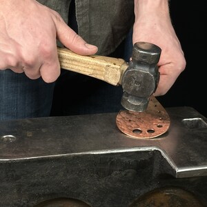 May include: A person using a hammer to shape a copper piece on an anvil.