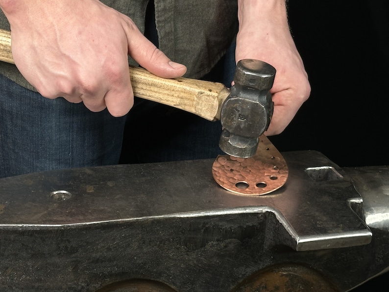 May include: A person using a hammer to shape a copper piece on an anvil.