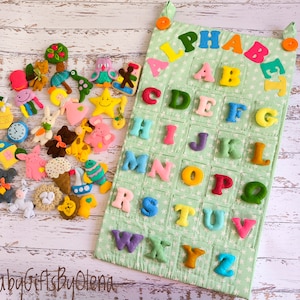 Könnte beinhalten: Ein Filz-Alphabet-Chart mit bunten Buchstaben und einer Sammlung von Filz-Tier- und Objektformen. Die Karte ist hellgrün mit weißen Sternen und hat eine Tasche für jeden Buchstaben. Die Formen umfassen einen Baum, einen Oktopus, einen Stern und ein Kaninchen.