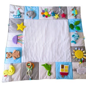 Può includere: Tappeto gioco per bambini con un pannello centrale bianco e un bordo patchwork. Il bordo include vari giocattoli morbidi come una barca, un elefante e una stella. I giocattoli sono di colori vivaci e realizzati con materiali morbidi.