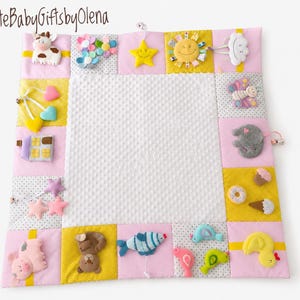 Pode incluir: Um tapete de atividades quadrado para bebé com um centro branco com relevo e uma borda de quadrados de tecido rosa, amarelo e cinzento com bolinhas. A borda apresenta vários apliques de peluche, incluindo uma vaca, estrela, sol e pato.