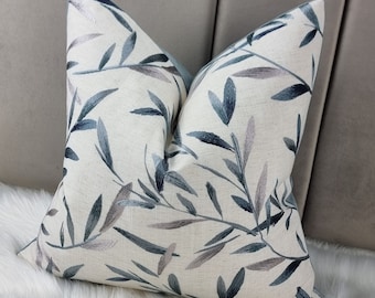 May include: A white cushion with an embroidered design of blue and grey leaves. The cushion is on a white fur surface.