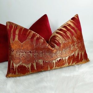 Peut inclure: Deux coussins décoratifs rouges et dorés. Le coussin avant présente un motif abstrait texturé or et rouge. Un coussin rouge uni est derrière. Les coussins sont sur une surface blanche et moelleuse.