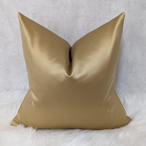 May include: A square, gold cushion with a textured surface. The cushion has a pointed top and is resting on a white, fluffy surface. The cushion is a solid colour and appears to be made of a soft material.