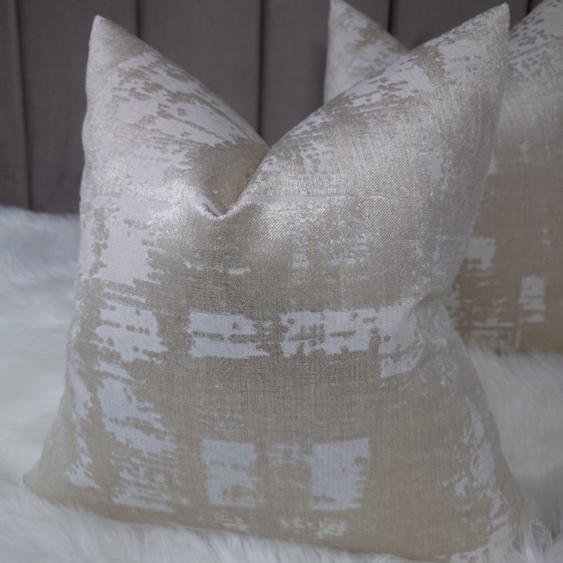 Puede incluir: Dos almohadas decorativas con un patr&oacute;n geom&eacute;trico blanco y dorado. Las almohadas est&aacute;n hechas de una tela suave y texturizada.