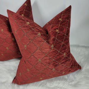 May include: Two red velvet cushion covers with a gold embroidered diamond pattern. The cushions are on a white fluffy surface.