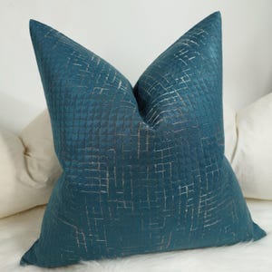 May include: Teal-colored decorative pillow with a geometric pattern. The pillow has a textured surface with a grid-like design in a lighter metallic color. The pillow is square-shaped and appears to be made of a soft fabric.