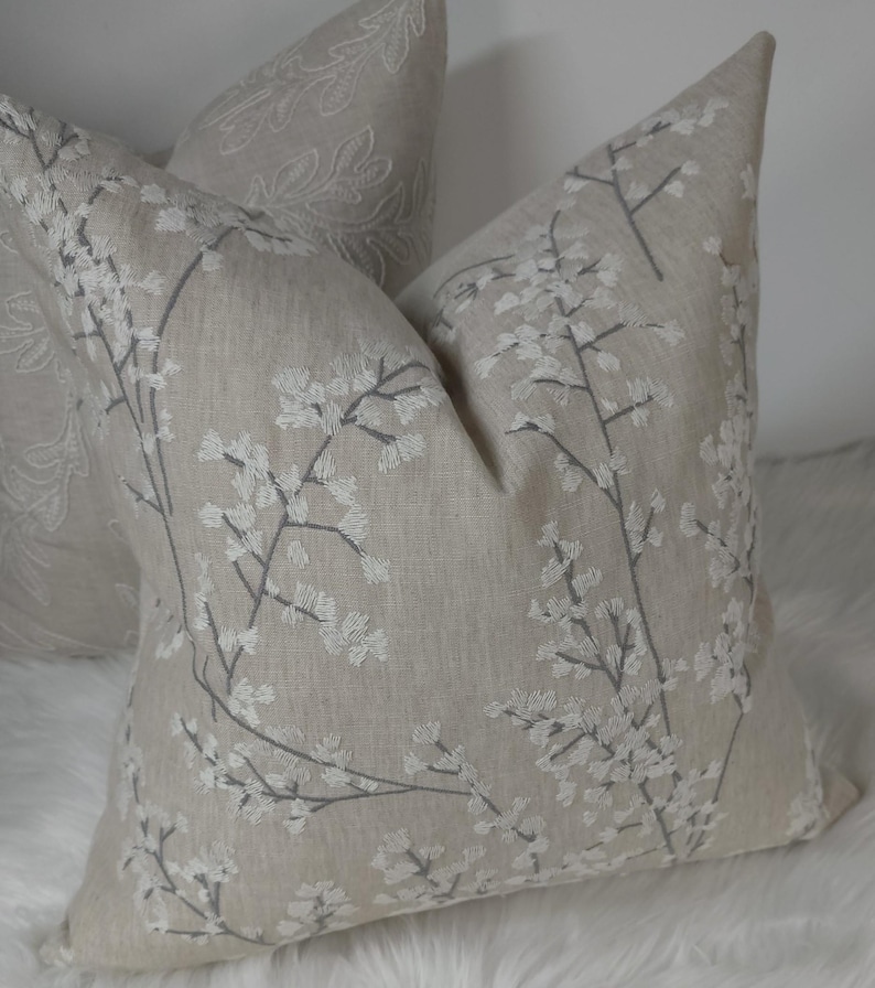 Peut inclure: Deux coussins en lin beige avec des motifs floraux brod&eacute;s. Le coussin de gauche a un motif plus complexe, tandis que le coussin de droite a un motif floral simple.