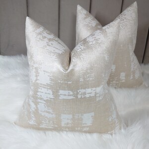 Puede incluir: Dos almohadas decorativas con un patr&oacute;n abstracto blanco y dorado. Las almohadas est&aacute;n hechas de una tela suave y texturizada.