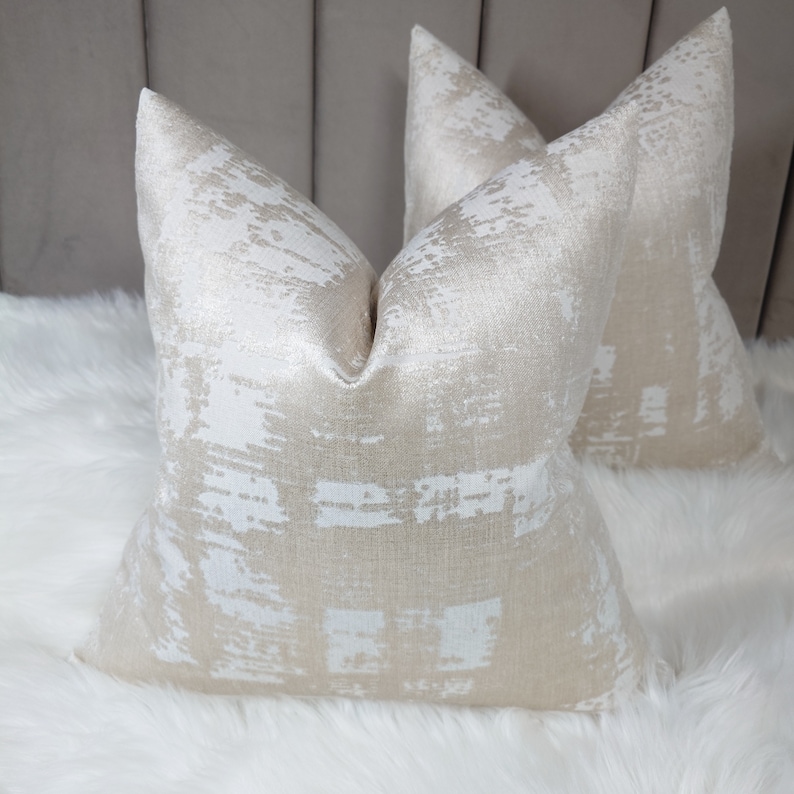 Puede incluir: Dos almohadas decorativas con un patr&oacute;n abstracto blanco y dorado. Las almohadas est&aacute;n hechas de una tela suave y texturizada.