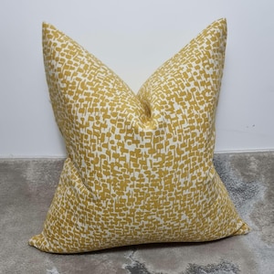 May include: A square throw pillow featuring a mustard yellow and white abstract block print pattern. The fabric displays a repeating design of small, irregular rectangular shapes arranged in a dense, textured layout across the entire surface.