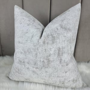 Può includere: Un cuscino bianco morbido con un sottile motivo grigio e bianco. Il cuscino è realizzato in tessuto strutturato e ha una forma quadrata.