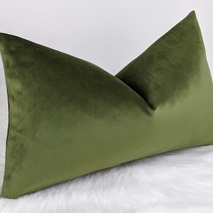 May include: Olive green velvet cushion. The rectangular cushion is a solid colour and has a soft, plush appearance. The cushion is placed on a white, fluffy surface.