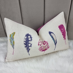 May include: A long rectangular throw pillow with a white linen fabric cover. The pillow features a colorful abstract design of five feathers in shades of pink, purple, blue, green, and yellow.