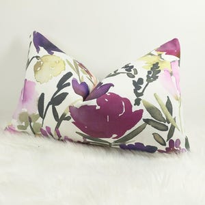 May include: Decorative pillow with a watercolor floral design. The pillow features large, deep purple and pink flowers with green and black foliage against a white background. The pillow is rectangular and has a soft, textured appearance.
