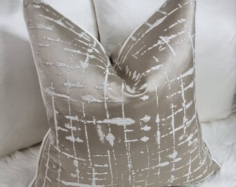 May include: A decorative throw pillow with a taupe background and a white, abstract grid pattern. The pillow has a white border and is placed in front of other white pillows. The pillow is square and has a soft, textured appearance.