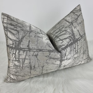 May include: Two decorative throw pillows with a textured, marbled design. The pillows are primarily light gray with darker gray vein-like patterns. The pillows are placed on a white, fluffy surface.
