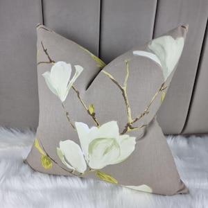 Könnte beinhalten: Ein dekoratives Kissen mit beigefarbenem Hintergrund und einem floralen Muster aus weißen Magnolienblüten mit gelben Akzenten.