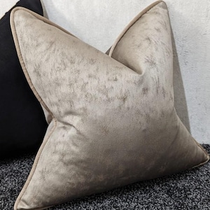 May include: A decorative pillow with a textured, light beige surface and a contrasting darker beige trim. The pillow has a soft, velvety appearance and is placed next to a black pillow. The pillow is likely for home decor.