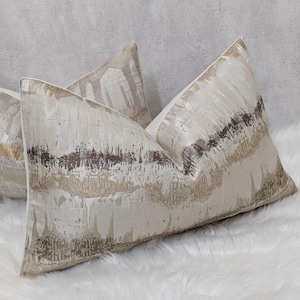 May include: Rectangular decorative cushions with a textured, abstract pattern in cream, beige, and brown hues. The cushions are arranged on a white, fluffy surface. The fabric has a subtle sheen. The design evokes a sense of luxury.