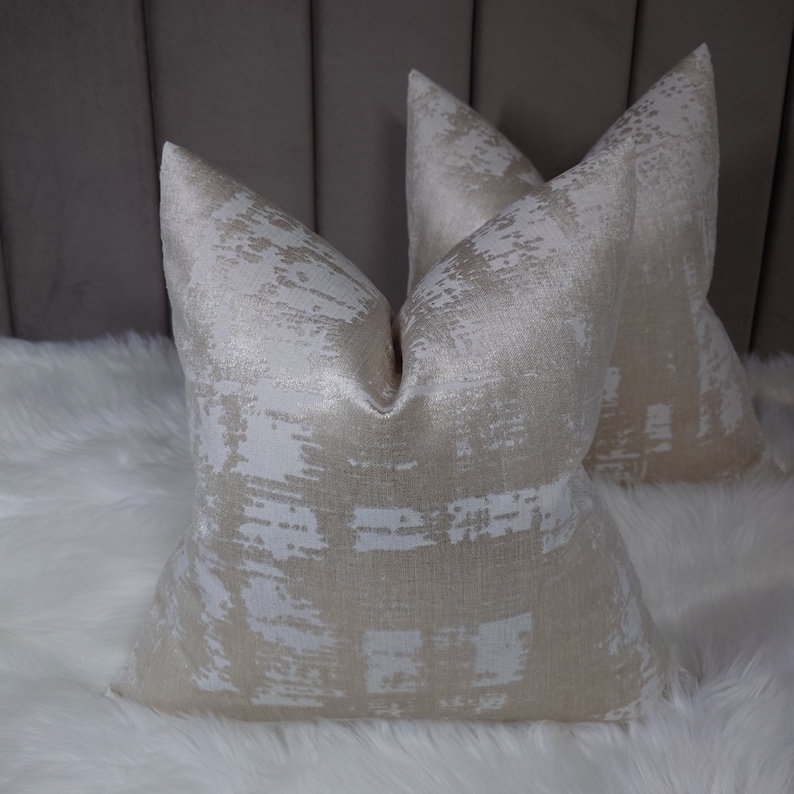 Puede incluir: Dos almohadas decorativas con un patr&oacute;n abstracto blanco y dorado. Las almohadas est&aacute;n hechas de una tela suave y texturizada y tienen un brillo sutil.