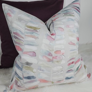 Puede incluir: Cojín decorativo con un patrón de acuarela en tonos rosa, azul, verde y blanco sobre un fondo gris claro. El cojín presenta un diseño repetido de formas abstractas. Detrás hay un cojín morado oscuro.