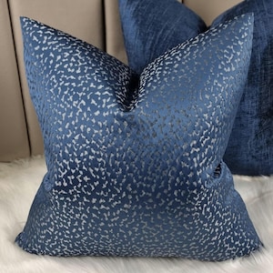 May include: Two navy blue cushions with a textured, speckled pattern. They rest on a white faux fur surface, with a neutral backdrop. The cushions have a soft, inviting appearance, perfect for home decor.