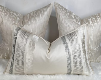 May include: Four decorative pillows in a neutral color palette. Two pillows have a textured, vertical stripe design. The other two pillows are rectangular with a cream background and a silver stripe pattern. The pillows are on a white, fluffy surface.