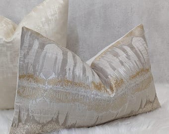 May include: Two decorative throw cushions. One is a creamy white colour with a subtle textured pattern. The other has a grey and gold abstract design. The cushions are on a white, fluffy surface.