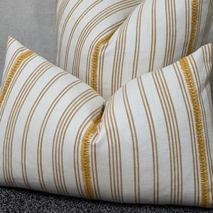 Puede incluir: Dos cojines decorativos con fondo blanco y rayas verticales color beige. Los cojines tienen un borde bordado amarillo. Los cojines están sobre una superficie gris oscuro.