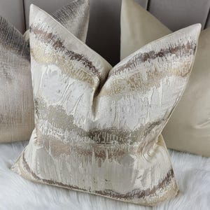 May include: A set of three decorative throw pillows with a cream background and abstract patterns in shades of gold, brown, and gray. The pillows are made of a soft, textured fabric.