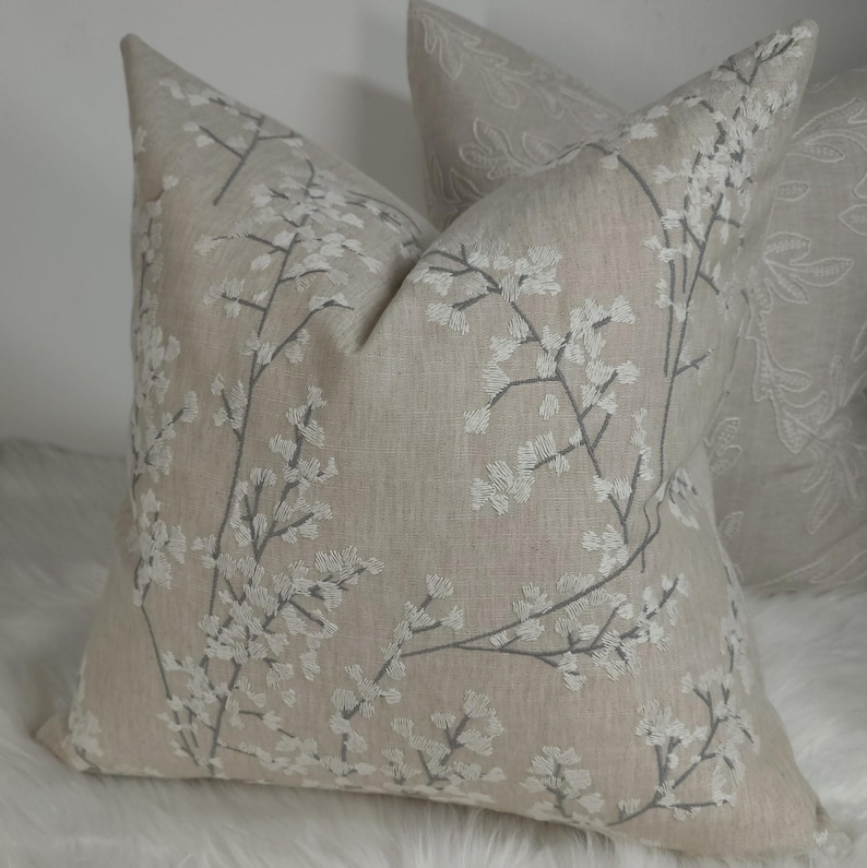 Peut inclure: Un oreiller en lin beige avec un motif floral brod&eacute; blanc. L'oreiller a un contour gris subtil sur le motif floral.