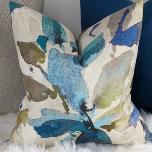 Op de afbeelding: Een decoratief kussen met een aquarelontwerp. Het kussen heeft een crèmekleurige achtergrond met blauwe, turquoise en bruine abstracte vormen. Het ligt op een witte, zachte ondergrond, met andere kussens op de achtergrond.