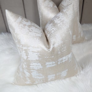 Puede incluir: Dos almohadas decorativas con un patr&oacute;n abstracto blanco y dorado. Las almohadas est&aacute;n hechas de una tela suave y texturizada.