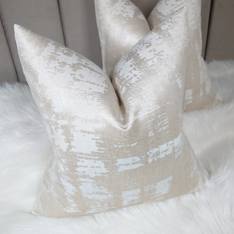 Puede incluir: Dos almohadas decorativas con un patr&oacute;n abstracto blanco y dorado. Las almohadas est&aacute;n hechas de una tela suave y texturizada.