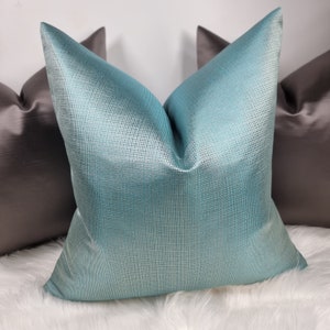 Op de afbeelding: Een tealblauw kussen met een subtiel geweven patroon. Het kussen ligt op een wit nep bont oppervlak.