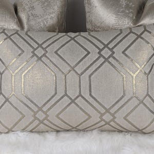 Puede incluir: Cojín rectangular decorativo con un patrón geométrico en plata y dorado. El cojín tiene un fondo texturizado blanquecino y un diseño repetido de líneas y formas que se cruzan. El cojín está colocado sobre una superficie blanca y esponjosa.