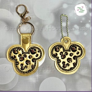 Könnte beinhalten: Zwei goldfarbene Schlüsselanhänger mit einem Leopardenmuster-Mickey-Mouse-Design. Die Schlüsselanhänger befinden sich auf einem weißen Hintergrund mit einem Glitzermuster. Der Text "Once Upon A Hoop Designs" ist im Hintergrund sichtbar.