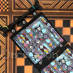 May include: A black and blue fabric wall hanging with a repeating pattern of Jack Skellington and Sally from the Nightmare Before Christmas. The fabric is sewn into a square shape and has a black border. The wall hanging is attached to a black metal frame with two Mickey Mouse ears.