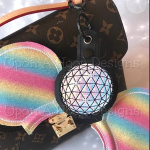 May include: Rainbow glitter Minnie Mouse ears with a black and silver sparkly ball charm attached to a black headband. The ears are on a brown and gold designer handbag.
