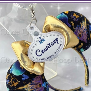 Pode incluir: Orelhas douradas de Minnie Mouse com um tecido preto e roxo com um castelo e silhuetas do Mickey Mouse. Uma etiqueta de nome branca com bordado dourado diz "Courtney" e "Where dreams come true".