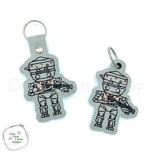 May include: Two embroidered keychains featuring a gray robot with a red and orange design. The robot is holding a gun and has a round head with red details. The keychains are on a white background.