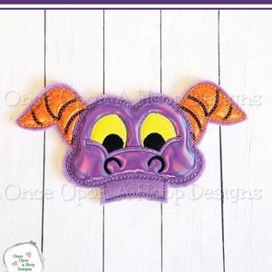 Op de afbeelding: Paarse en oranje vilt applique van een drakengezicht met grote gele ogen. De draak heeft zijn mond open en heeft een kleine roze neus.