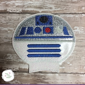 May include: A white and silver embroidered patch of a robot character with blue details. The robot has a round head with a red circle in the center and three horizontal lines below.