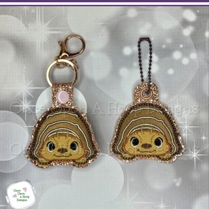 May include: Two keychains featuring a cartoon sloth with brown fur and a pink nose. The sloth is wearing a brown shell with gold glitter. One keychain has a gold chain and a gold clasp. The other keychain has a brown chain and a brown clasp.