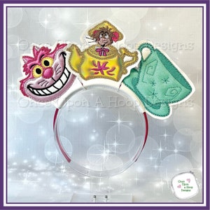 May include: A white headband with a pink Cheshire Cat, a yellow teapot with a mouse inside, and a green teacup. The headband is decorated with glitter and stars.