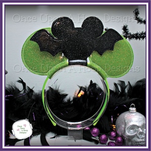 Puede incluir: Orejas de ratón de purpurina verde y negra con un diseño de murciélago. Las orejas están en una diadema. El texto "Once Upon a Hoop Designs" es visible en el fondo.