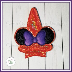 May include: Orange glitter witch hat with black ears and a purple bow. The hat is on a white wood background.